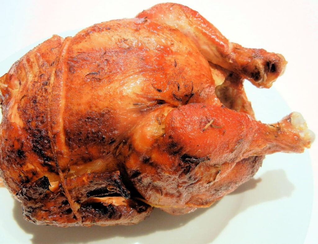 A whole golden-brown rotisserie chicken plated on white, showing tied legs and moist skin.