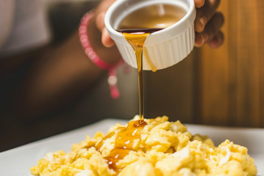 a ramekin of maple syrup being poured over scrambled eggs