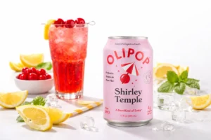 OLIPOP Shirley Temple soda can in the foreground with a sparkling Shirley Temple drink behind it, surrounded by cherries, lemon slices, ice, and mint on a clean white studio background.