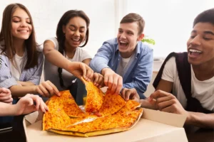 Teenagers grabbing pizza slices at a party while serving themselves from large boxes.