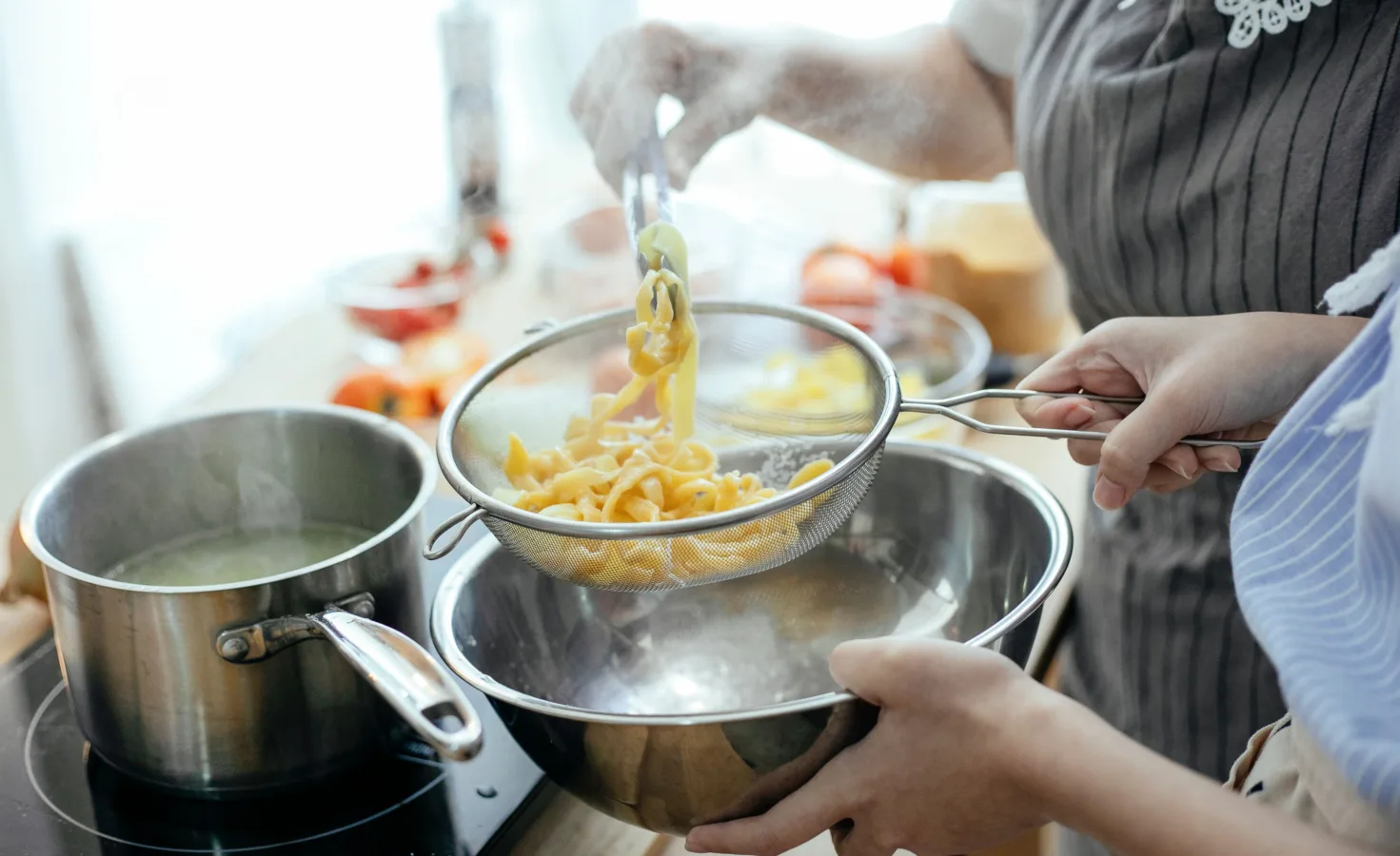 Two people draining freshly cooked pasta through a metal colander into a large bowl.